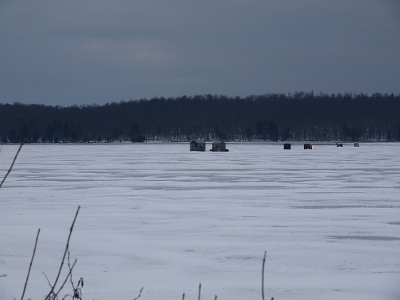[Different view of the same pond with 4 wooden shacks one car and one snowmobile on the ice.]
